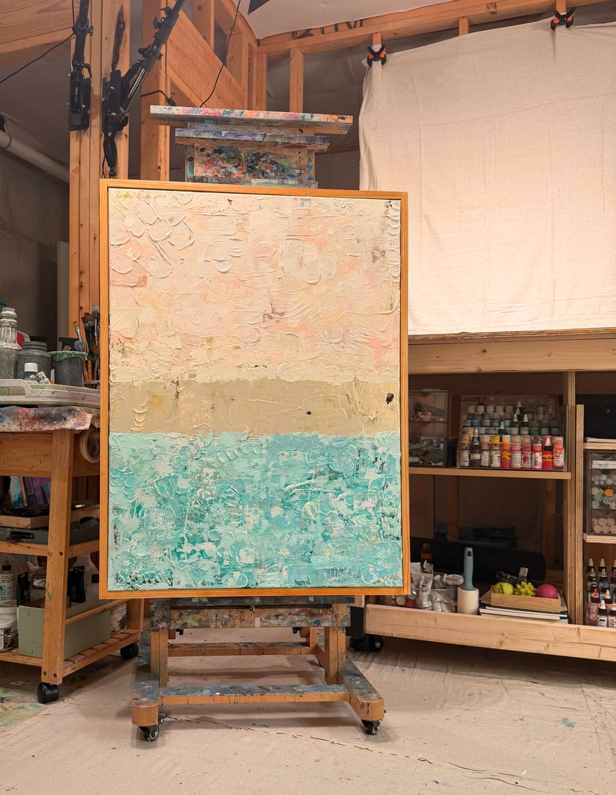 The Money Keeps Us Divided on an easel in Kindra's studio: textured blush pink above, a gold band across the middle, and speckled turquoise below.