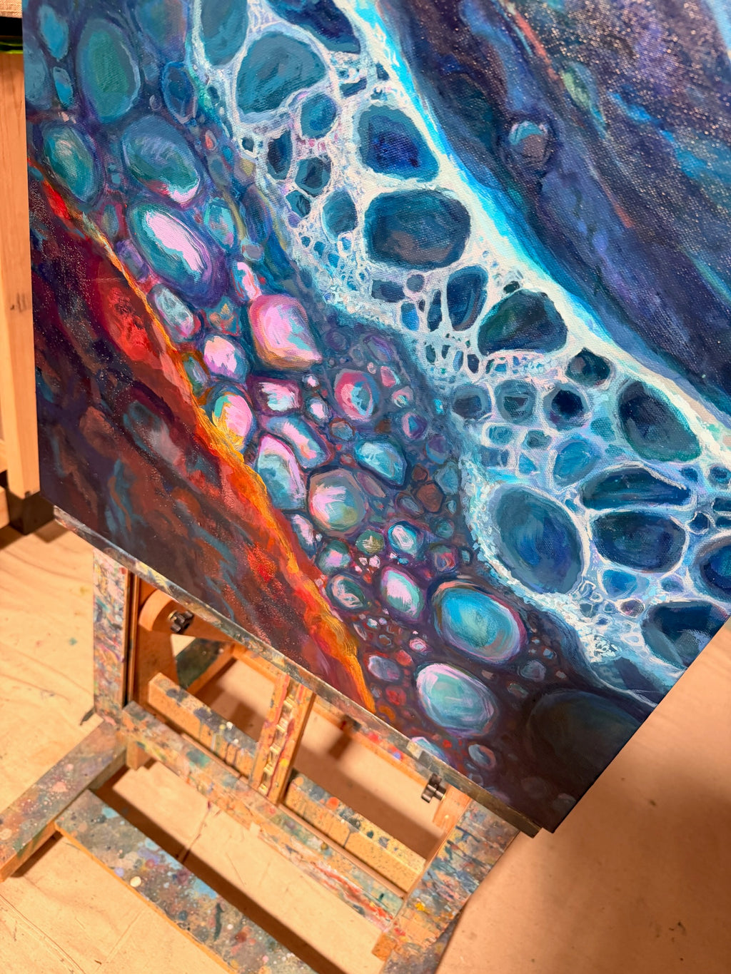 Angled close-up of Polished By The Rough Torrent on the easel, showing white foam-like lace around blue, pink and teal pebbles and a fiery orange edge.