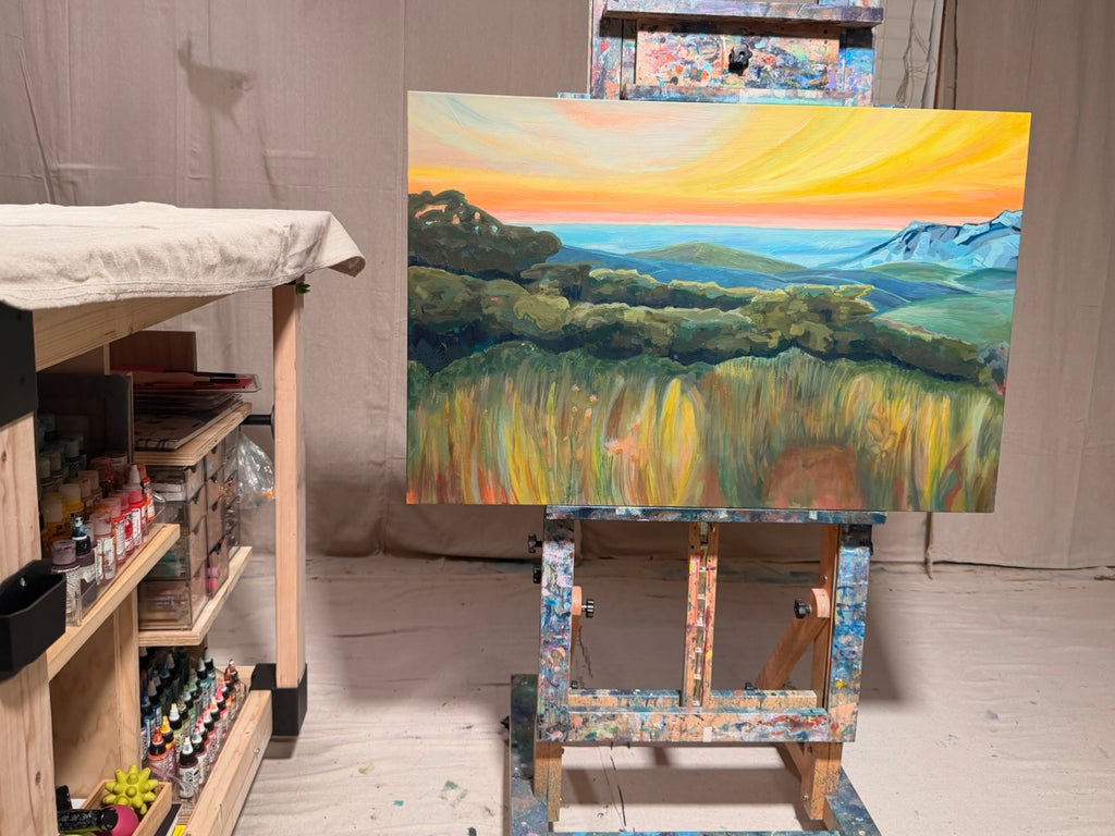 By Way Of The Weeds on a studio easel: an orange and yellow sunset sky over blue mountains, rolling green hills and a foreground of tall golden grass.