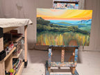 By Way Of The Weeds on a studio easel: an orange and yellow sunset sky over blue mountains, rolling green hills and a foreground of tall golden grass.