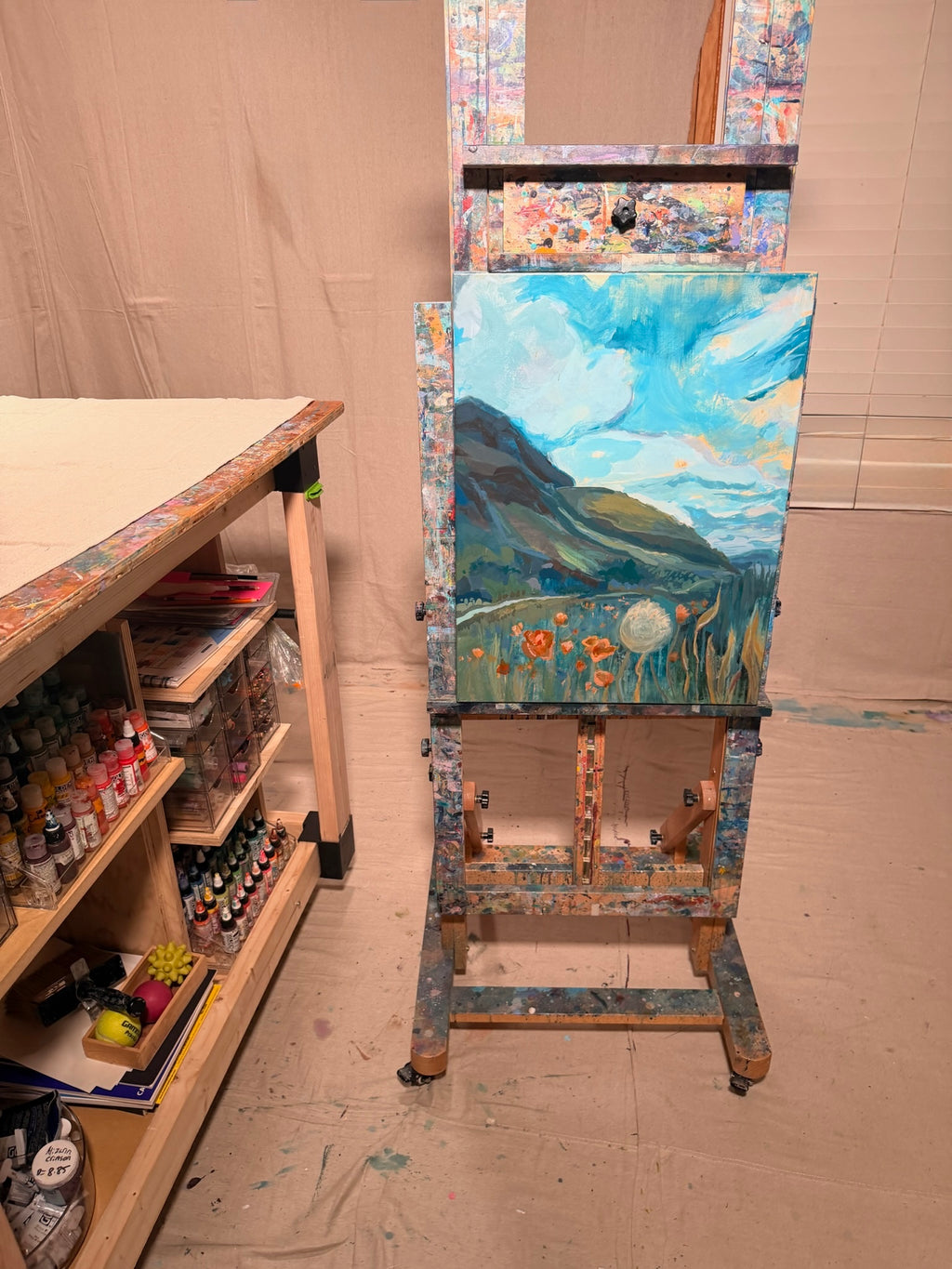 My Once Obliterated Self-Esteem resting on a paint-splattered easel in Kindra's studio: a green hill under a bright blue sky over a meadow of orange poppies and a dandelion.