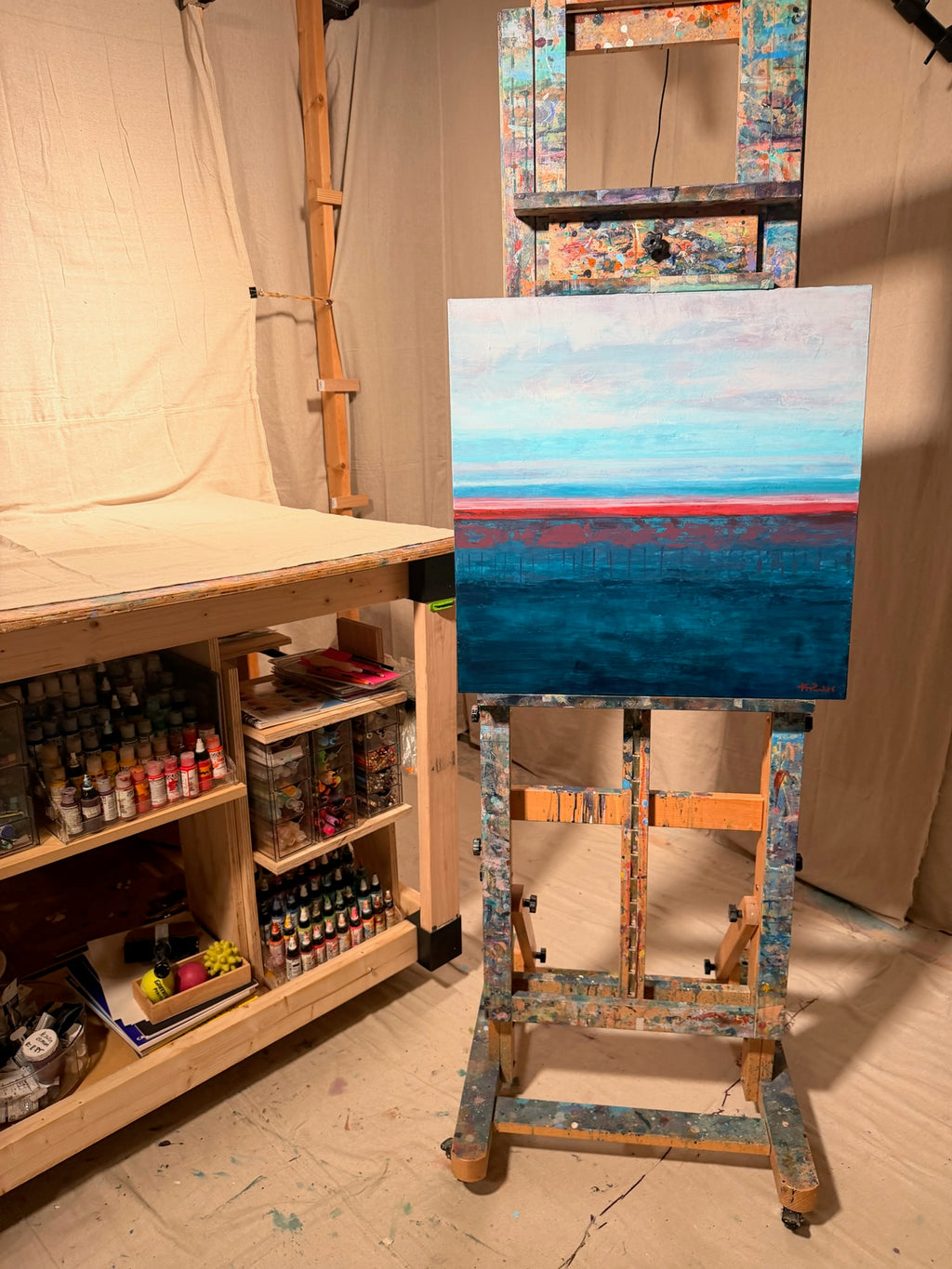Rest in Your Peace on an easel in Kindra's studio beside shelves of paint, showing a pale sky, coral horizon line and deep teal sea.