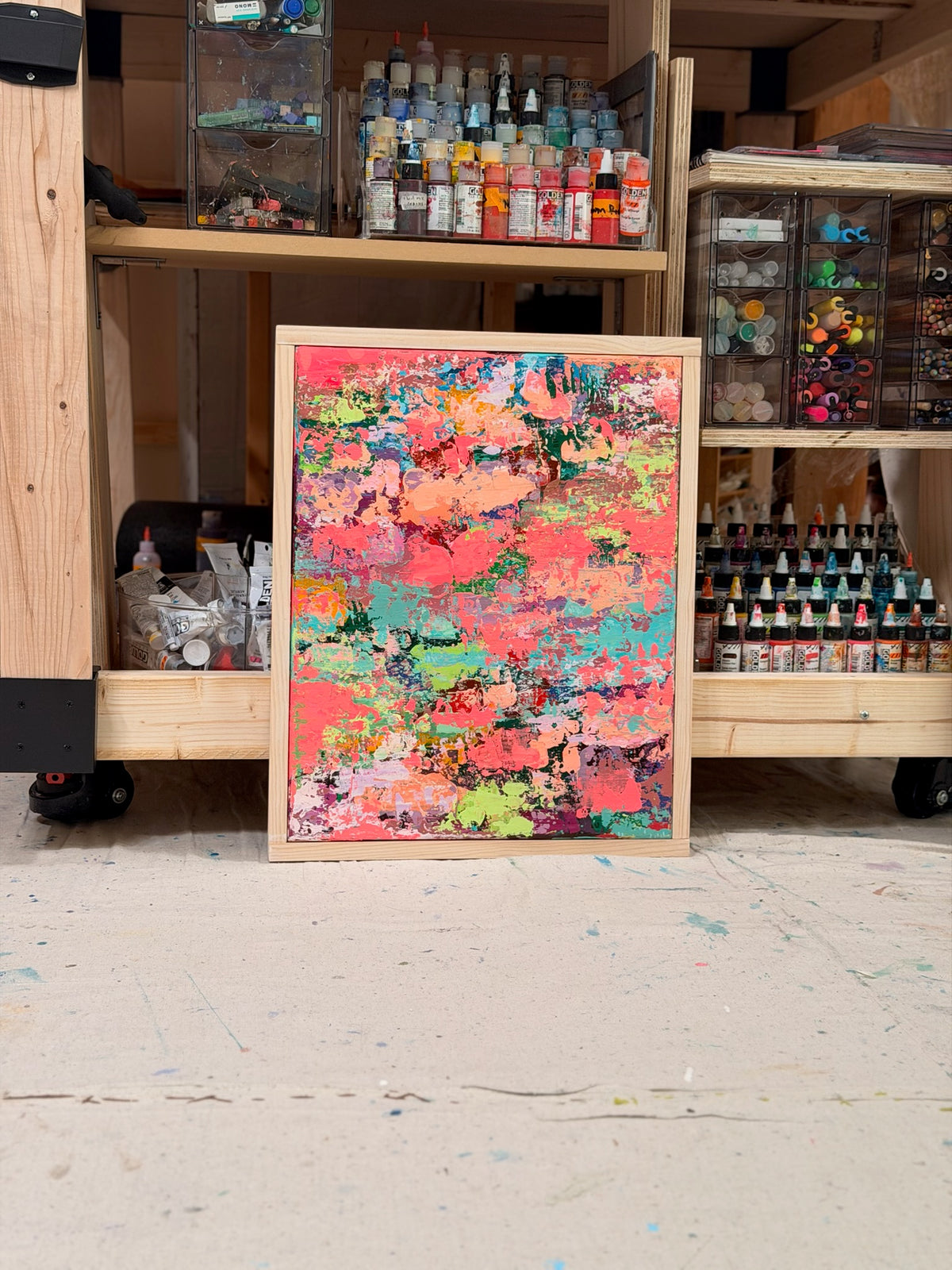 Pretty Girls Walk leaning against a studio supply shelf full of paints: a framed burst of coral, pink, teal and lime textured paint.