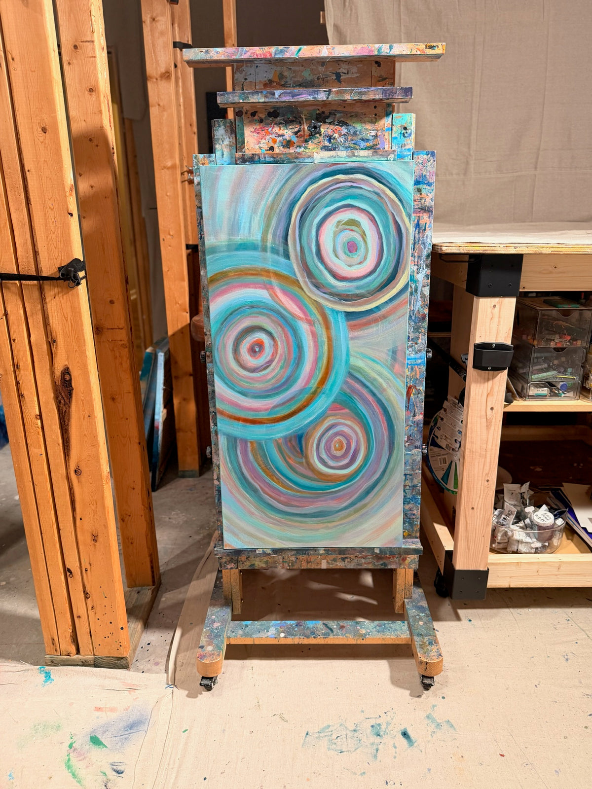 Abundance on a paint-splattered easel in Kindra's studio: three overlapping ripple circles in turquoise, pink, cream and copper on a tall canvas.