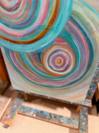 Angled close-up of Abundance on the easel, showing a pink-centered ripple framed by sweeping teal and copper rings.