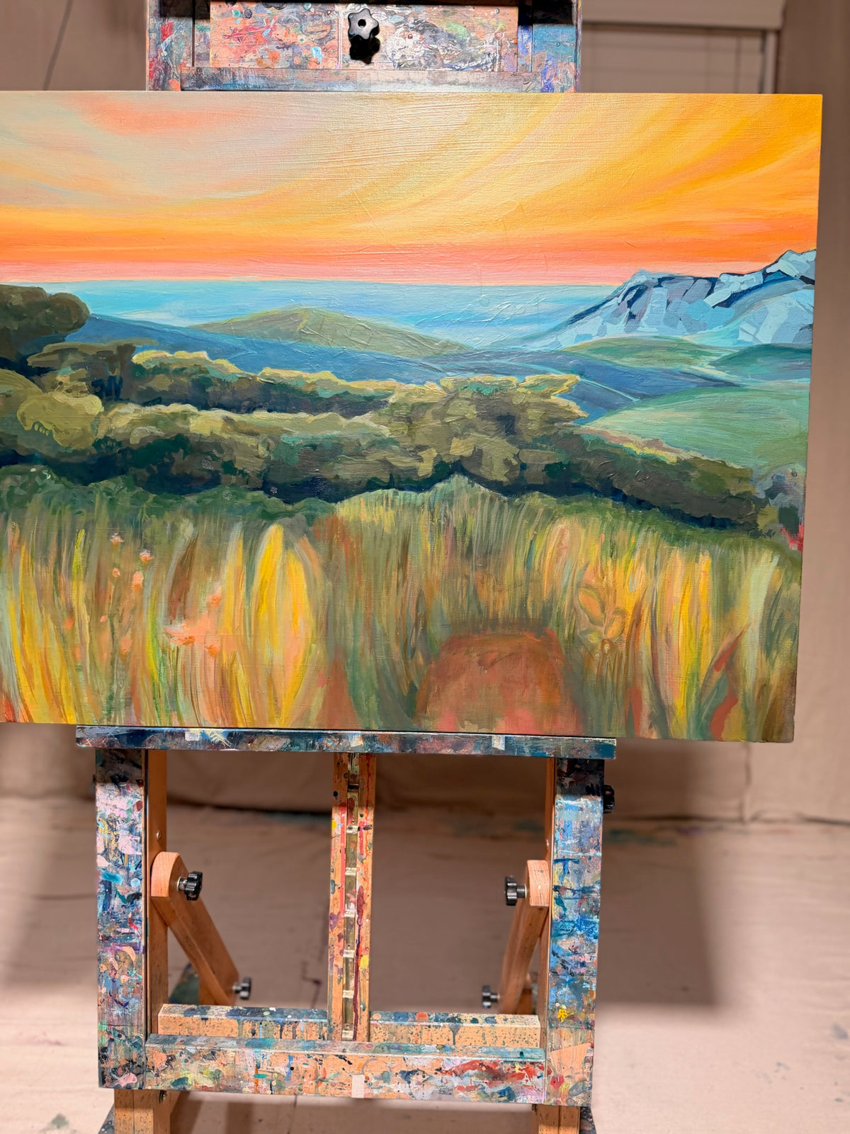 By Way Of The Weeds on a paint-splattered easel, viewed straight on: sunset sky, pale blue distant mountains, dark green trees and textured golden weeds.