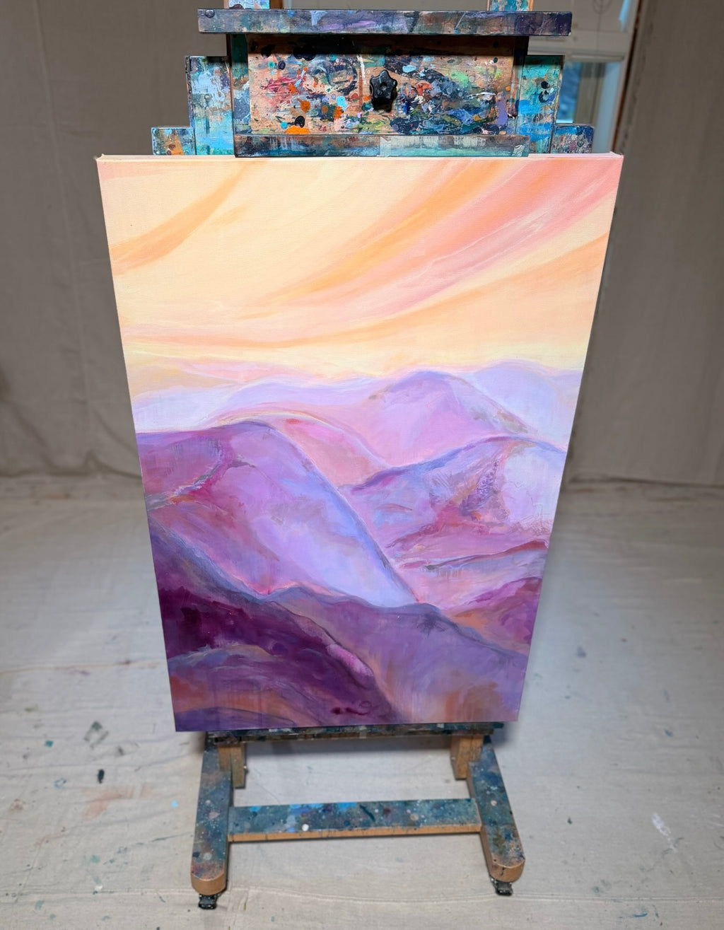 The View From My Latest Summit on a paint-splattered studio easel: sweeping peach and pink sky over rolling violet and magenta mountains.