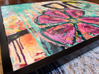 Angled close-up of In Bloom showing the textured pink flower, turquoise drips and black arches inside the black frame.