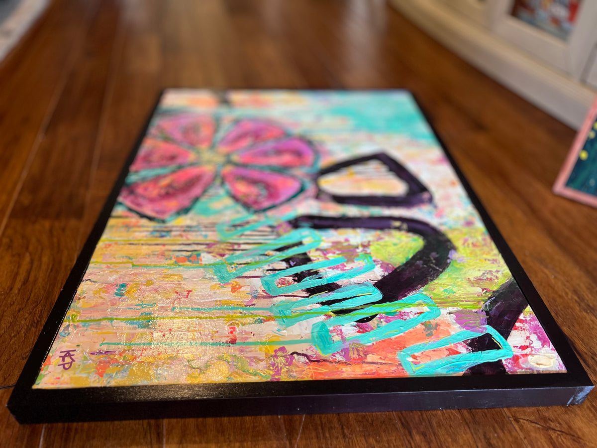 Low-angle view of In Bloom lying on a wood floor, showing its black frame, pink flower and turquoise scribbled marks.