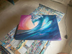 Held In The Current resting on a paint-splattered easel tray in the studio: a deep teal wave curls before a warm pink and peach sky.