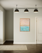The Money Keeps Us Divided shown on a pale wall in a hallway beneath black pendant lights, its blush, gold and turquoise bands framed in wood.