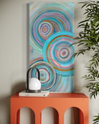 Abundance leaning above an orange arched side table with a lantern and books, its turquoise and pink ripple circles beside a leafy plant.