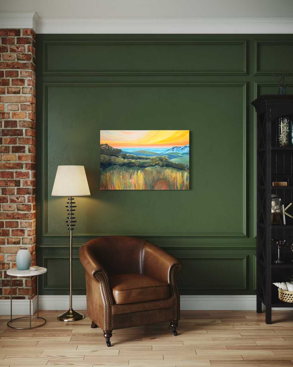 By Way Of The Weeds hanging on a dark green paneled wall above a brown leather armchair and floor lamp, its sunset glowing over golden fields.